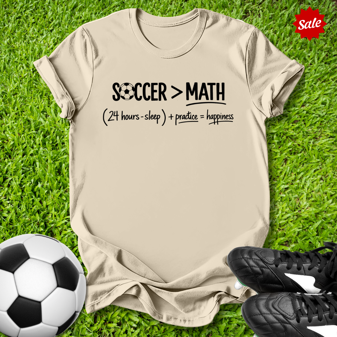 Beige t-shirt with 'Soccer > Math' text on grass with soccer ball and shoes
