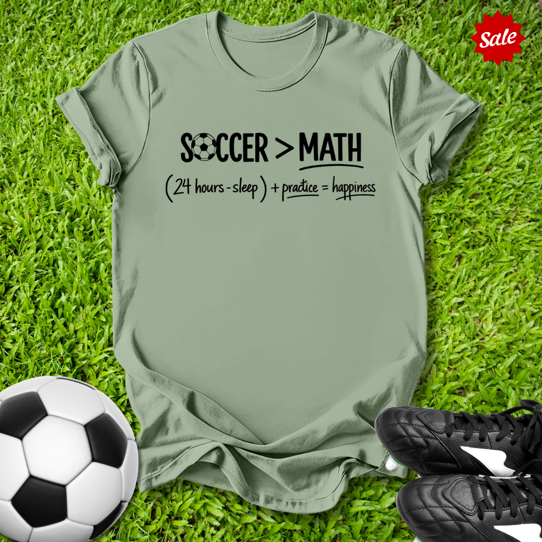Green t-shirt with 'Soccer > Math' text on grass with soccer ball and shoes