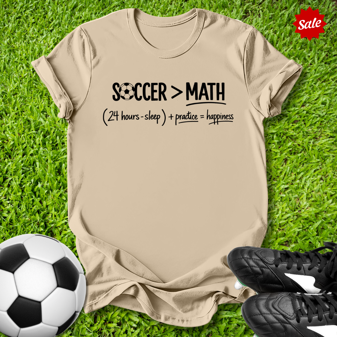Beige t-shirt with 'Soccer > Math' text on grass with soccer ball and shoes