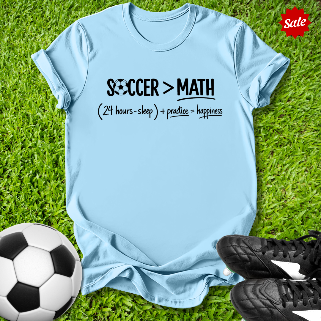 Blue t-shirt with 'Soccer > Math' text on grass with soccer ball and shoes