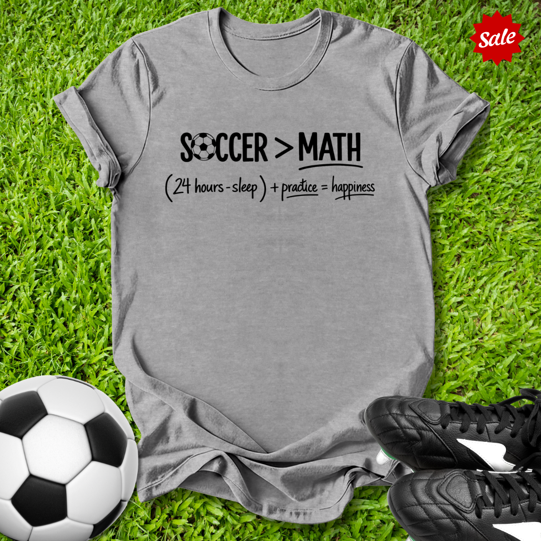 Gray t-shirt with 'Soccer > Math' text on grass with soccer ball and shoes