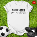 White t-shirt with 'Soccer > Math' text on grass with soccer ball and shoes