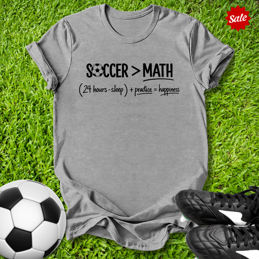Gray t-shirt with 'Soccer > Math' text on grass with soccer ball and shoes
