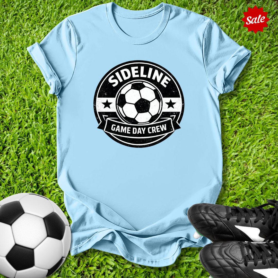 Light blue t-shirt with soccer-themed design on grass with soccer ball and shoes