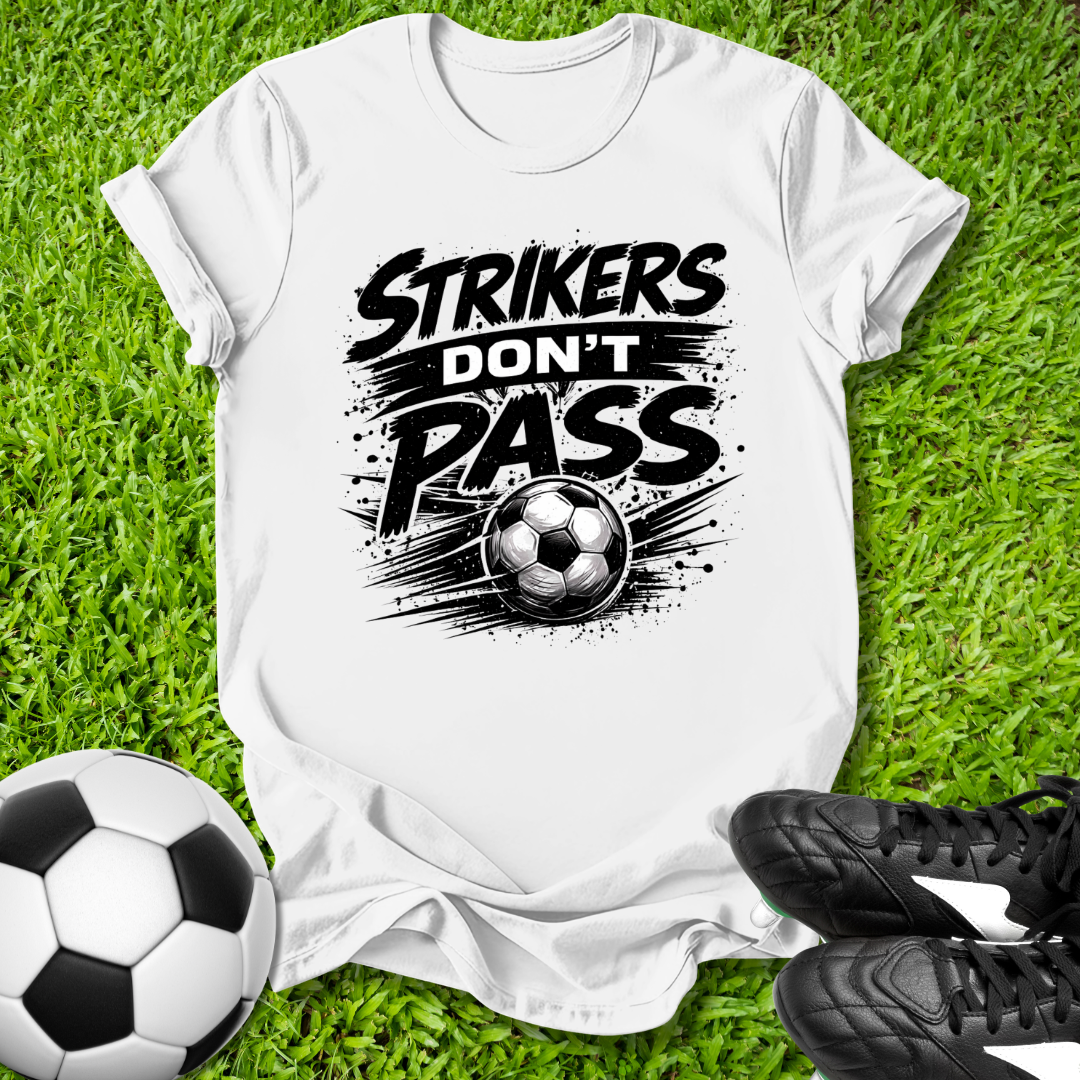 White t-shirt with 'Strikers Don't Pass' graphic on grass with soccer ball and cleats.