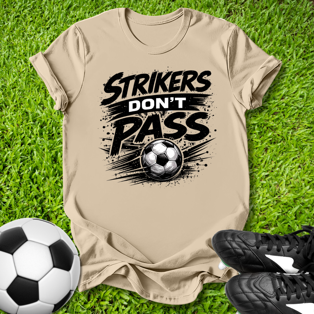 Beige t-shirt with 'Strikers Don't Pass' graphic on grass with soccer ball and shoes.