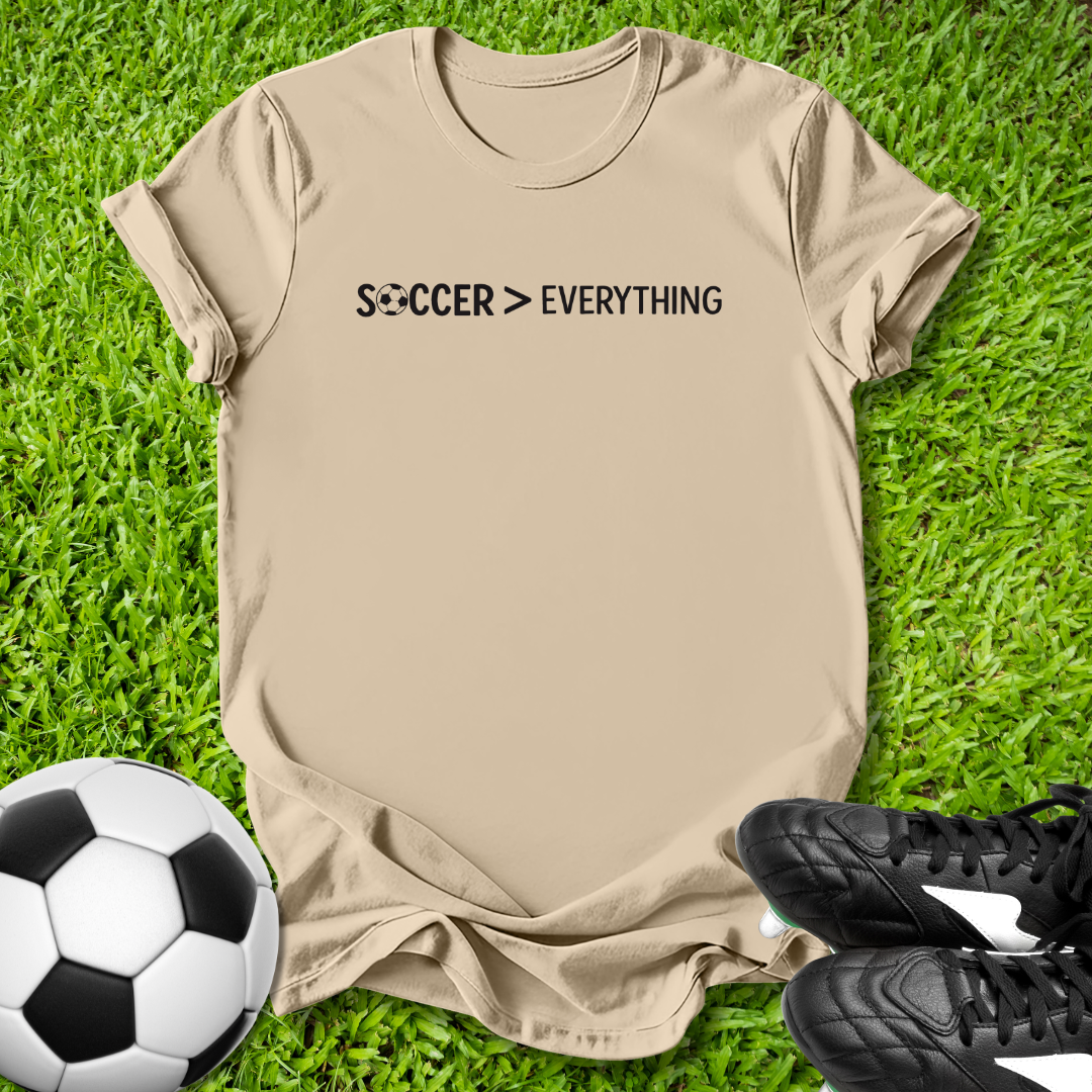 Beige t-shirt with 'Soccer > Everything' text on grass with soccer ball and shoes.