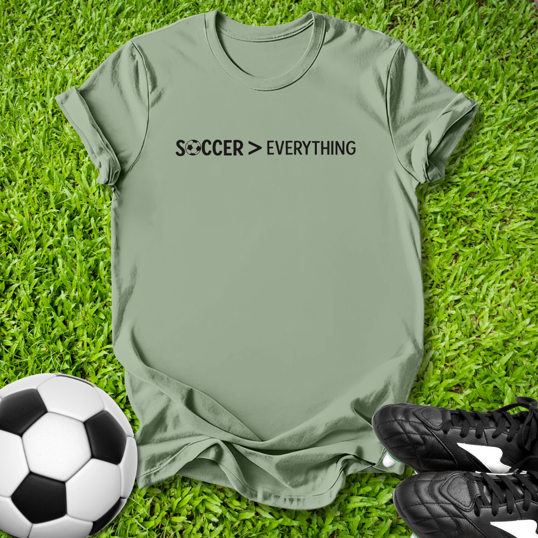 Green t-shirt with 'Soccer > Everything' text on grass with soccer ball and shoes.