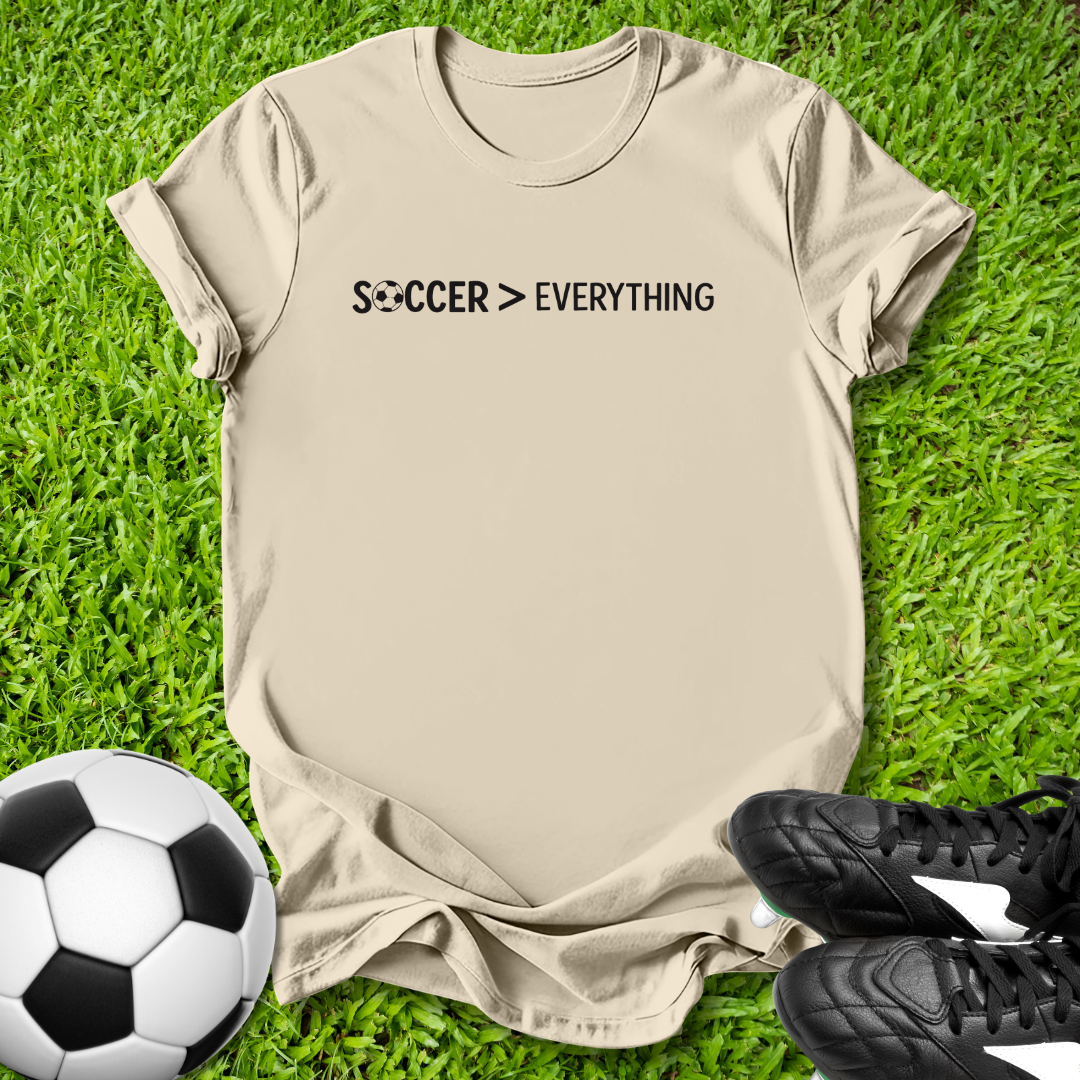 Beige t-shirt with 'Soccer > Everything' text on grass with soccer ball and shoes.