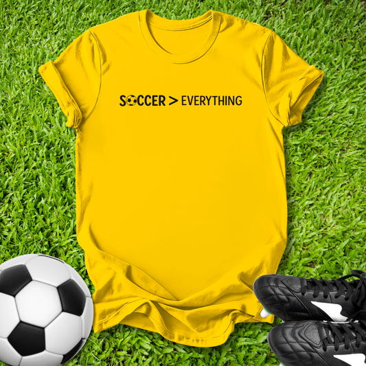 Yellow t-shirt with 'SOCCER > EVERYTHING' text on grass with soccer ball and shoes.