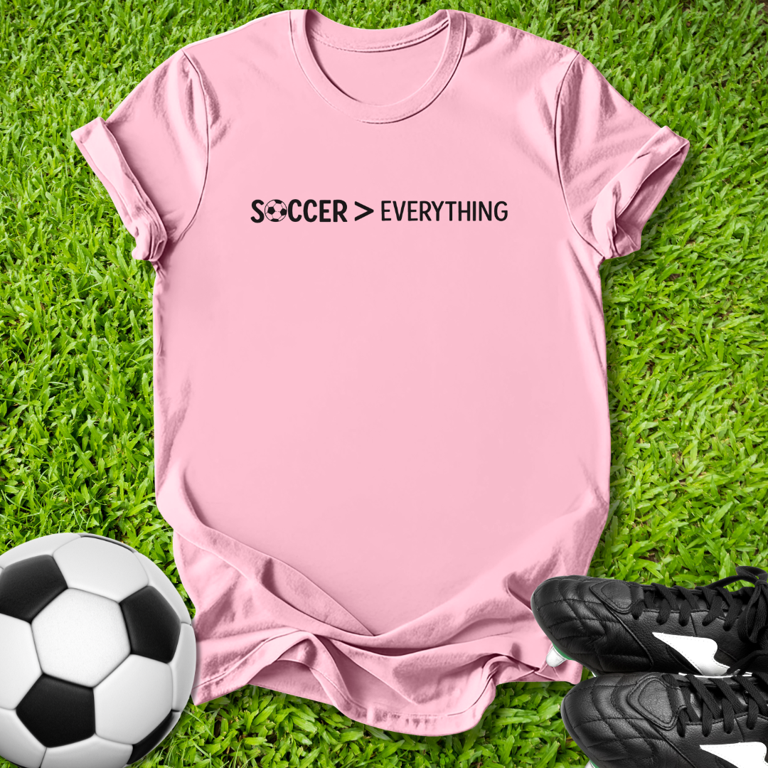 Pink t-shirt with 'Soccer > Everything' text on grass with soccer ball and shoes.