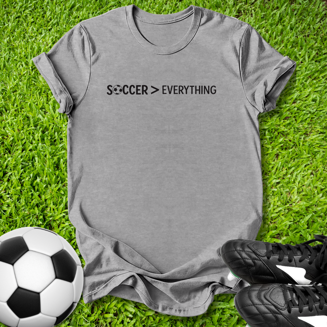 Gray t-shirt with 'Soccer > Everything' text on grass with soccer ball and shoes.