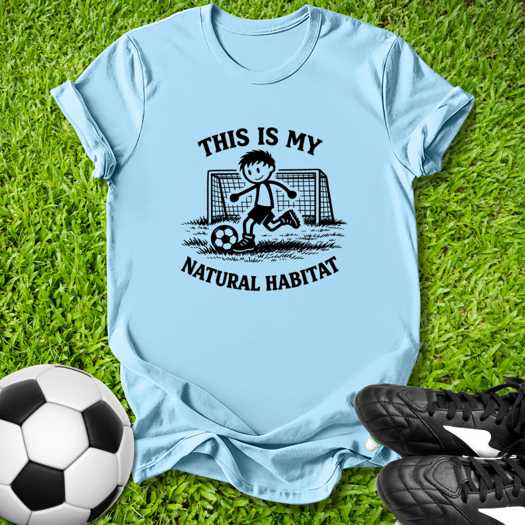 Light blue t-shirt with soccer graphic and this is my natural habitat text on grass with soccer ball and shoes.