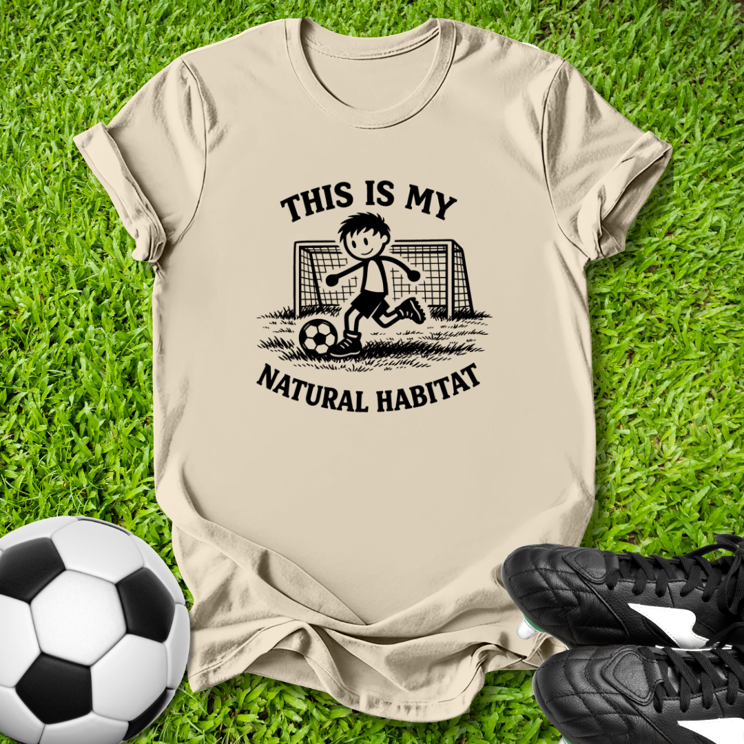 Beige t-shirt with soccer graphic and  this is my natural habitat text on grass with soccer ball and shoes.