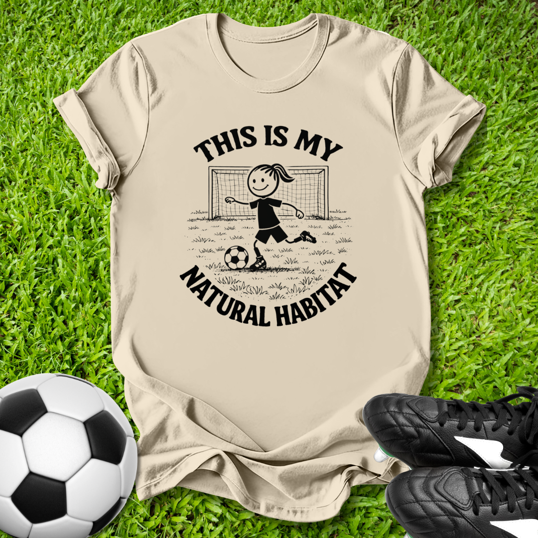 Beige t-shirt with soccer-themed  this is my natural habitat graphic and text on grass with soccer ball and shoes.
