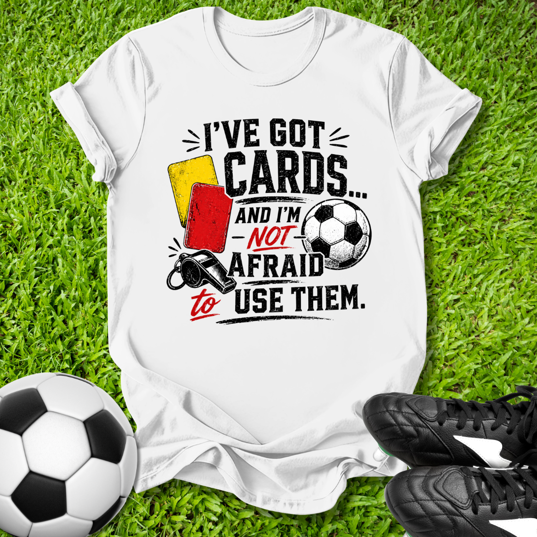 White t-shirt with soccer-themed text and graphics on grass with soccer ball and shoes.