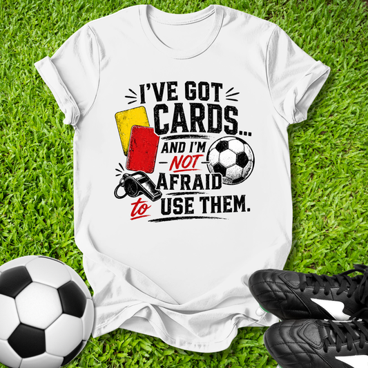 White t-shirt with soccer-themed text and graphics on grass with soccer ball and shoes.