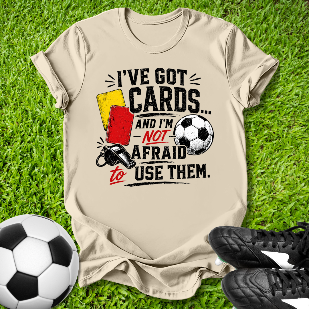 Beige t-shirt with soccer-themed text and graphics on grass with soccer ball and shoes.
