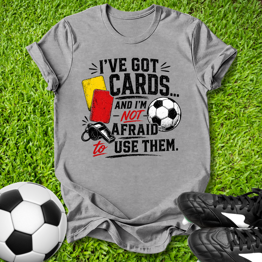 Gray t-shirt with soccer-themed text on grass with soccer ball and shoes