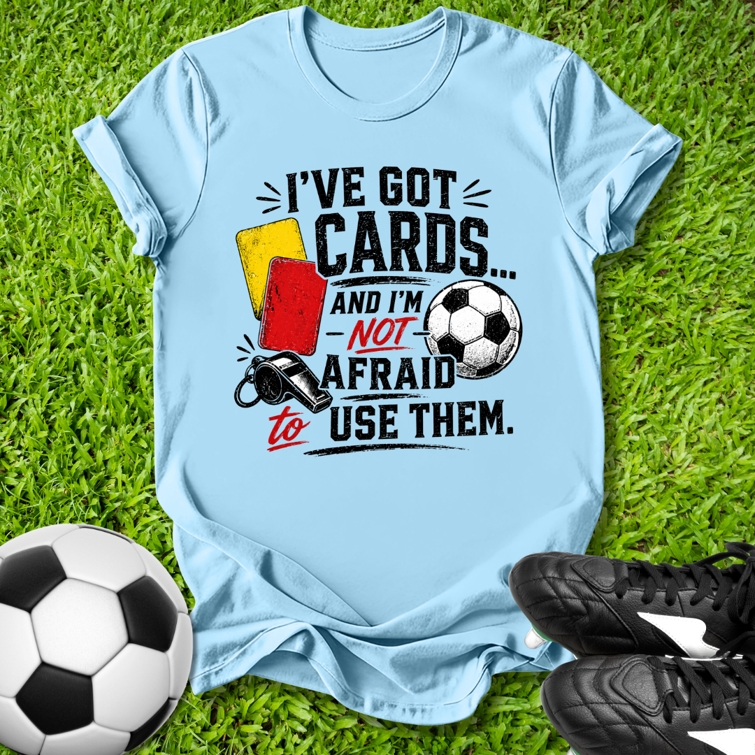 Light blue t-shirt with soccer-themed graphic and text on grass with soccer ball and shoes.