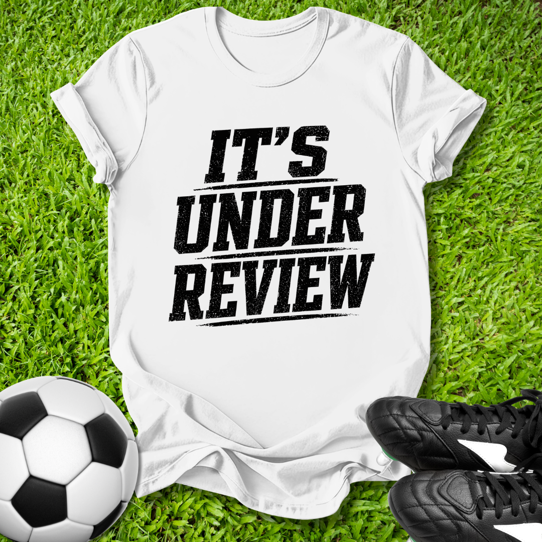White t-shirt with 'IT'S UNDER REVIEW' text on grass with soccer ball and shoes