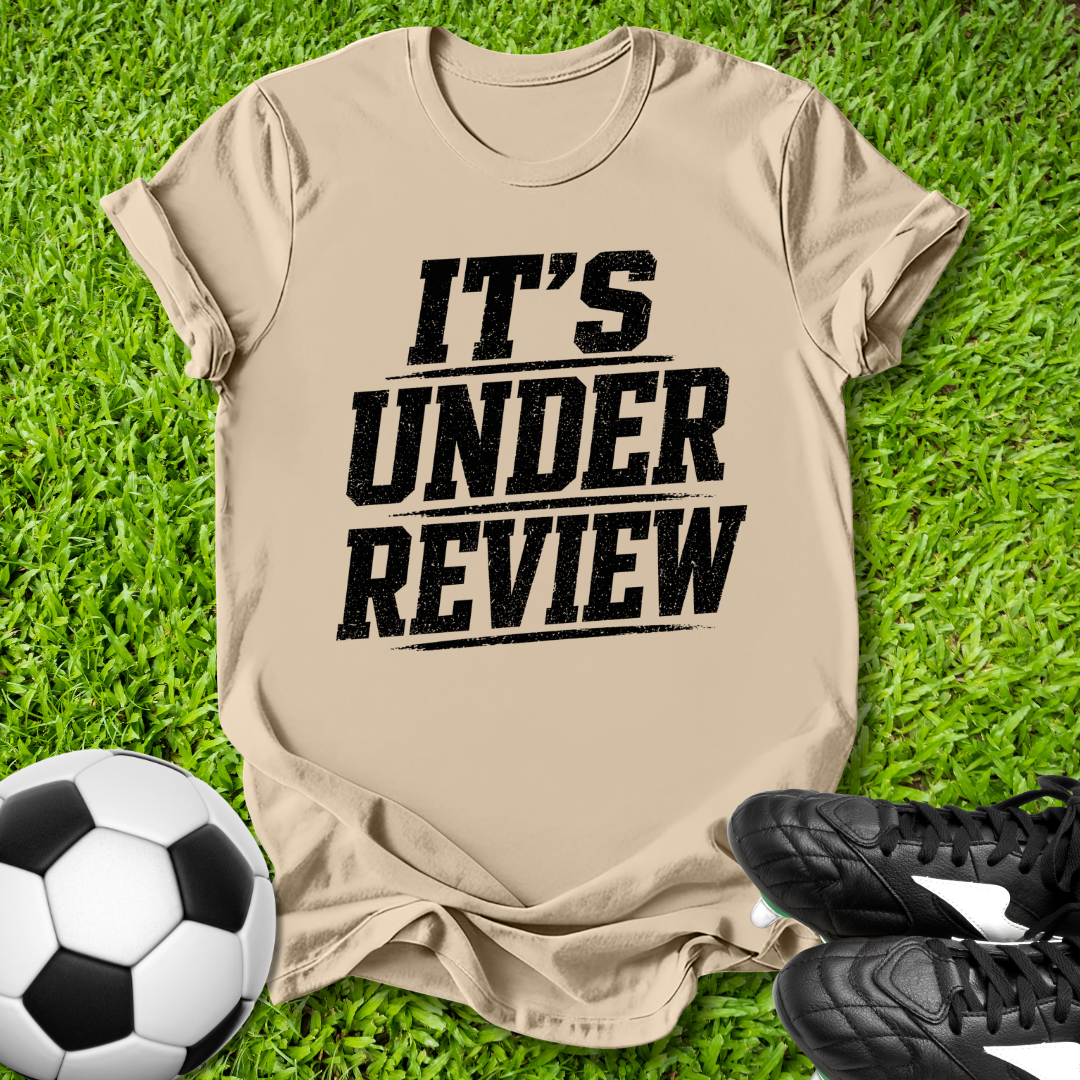Beige t-shirt with 'IT'S UNDER REVIEW' text on grass with soccer ball and shoes.