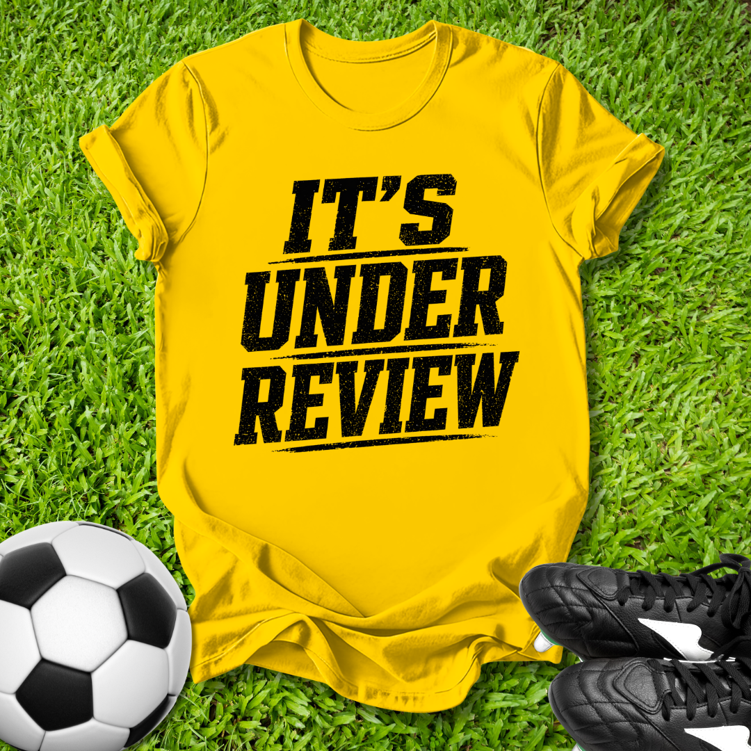 Yellow t-shirt with 'IT'S UNDER REVIEW' text on grass with soccer ball and shoes.