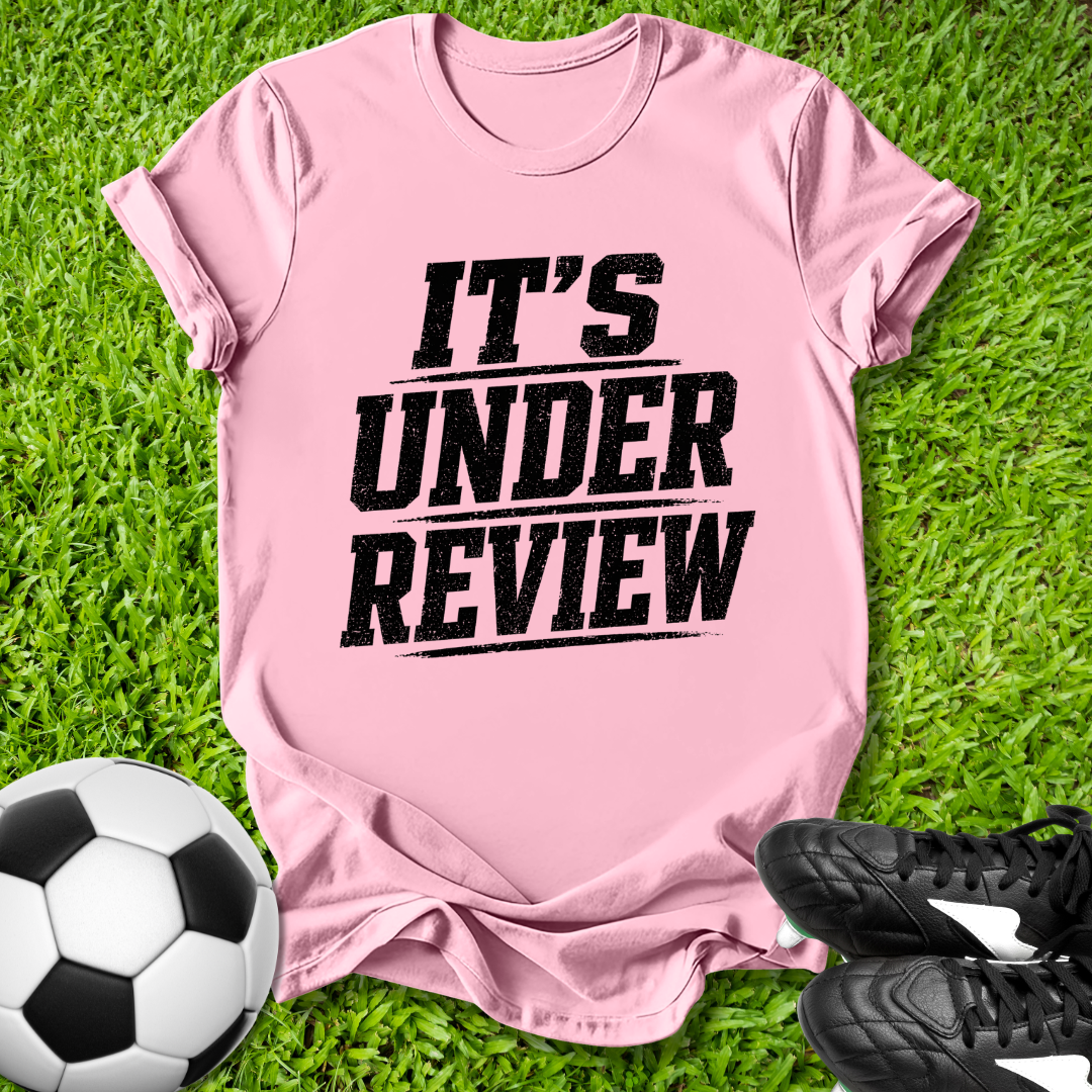 Pink t-shirt with 'IT'S UNDER REVIEW' text on grass with soccer ball and shoes