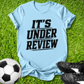 Light blue t-shirt with 'IT'S UNDER REVIEW' text on grass with soccer ball and shoes.