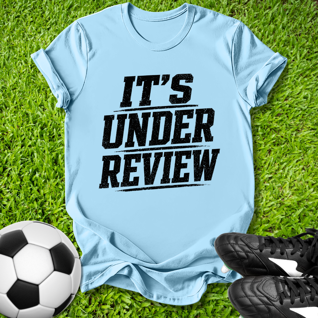 Light blue t-shirt with 'IT'S UNDER REVIEW' text on grass with soccer ball and shoes.