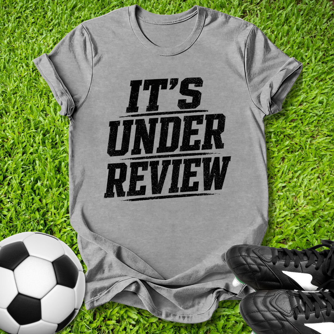 Gray t-shirt with 'IT'S UNDER REVIEW' text on grass with soccer ball and shoes