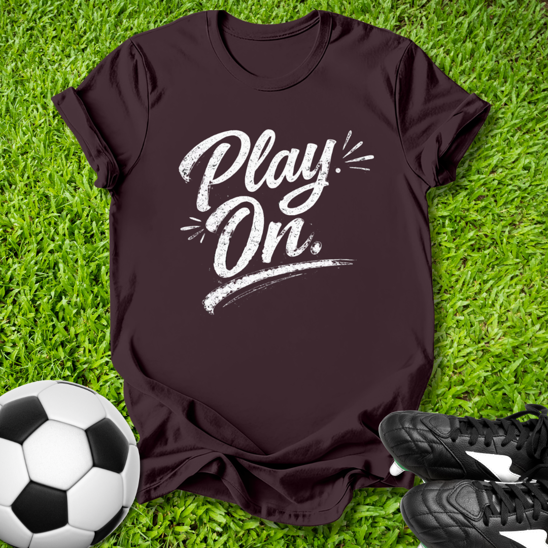 Brown t-shirt with 'Play On' text on grass with soccer ball and shoes