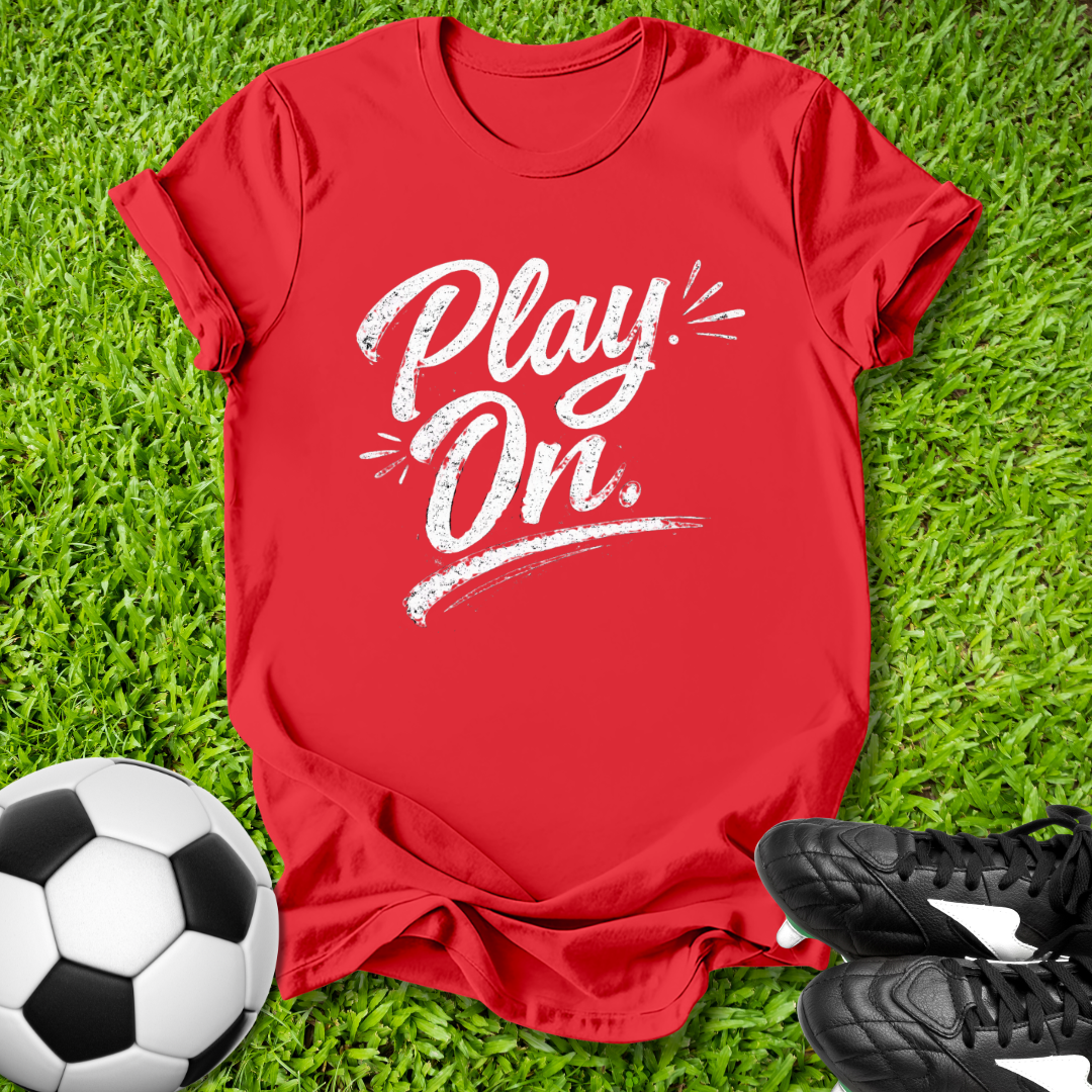 Red t-shirt with 'Play On' text on grass with soccer ball and shoes