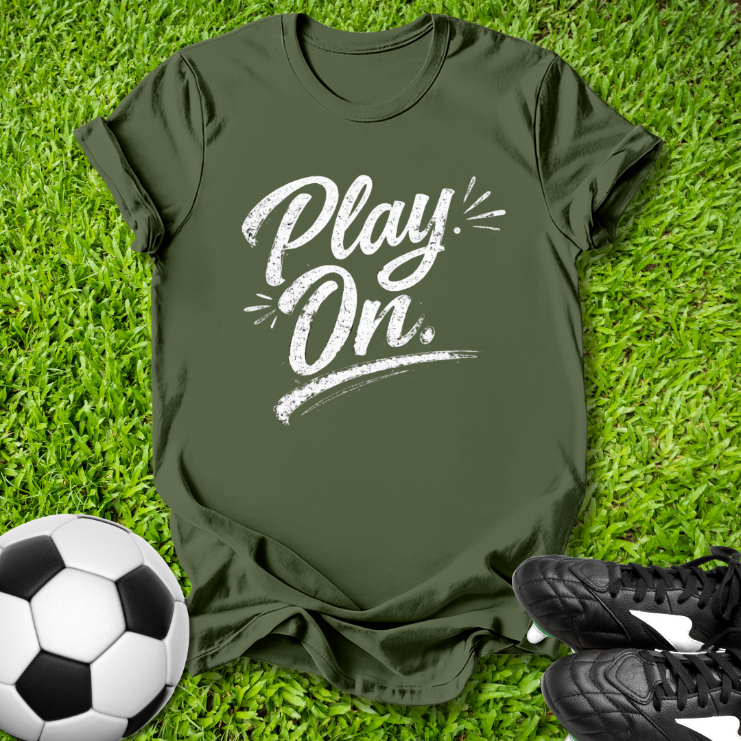 Green t-shirt with 'Play On' text on grass with soccer ball and shoes