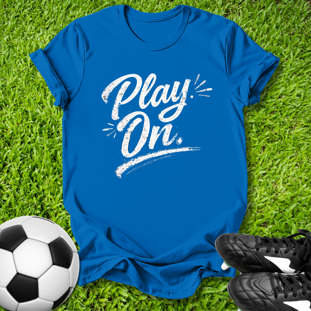 Blue t-shirt with 'Play On' text on grass with soccer ball and shoes