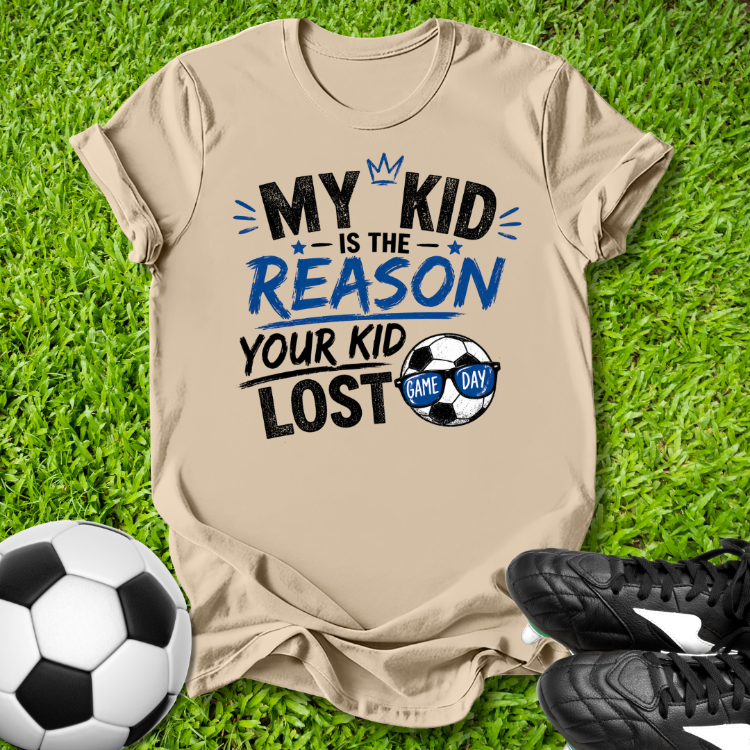 Beige t-shirt with humorous My Kid is the Reason Your Kid Lost text soccer-themed text on grass with soccer ball and shoes.