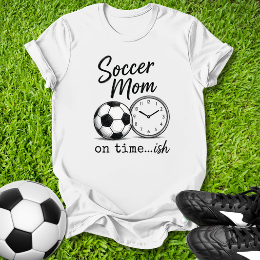 White t-shirt with 'Soccer Mom on time...ish' design on grass with soccer ball and cleats.