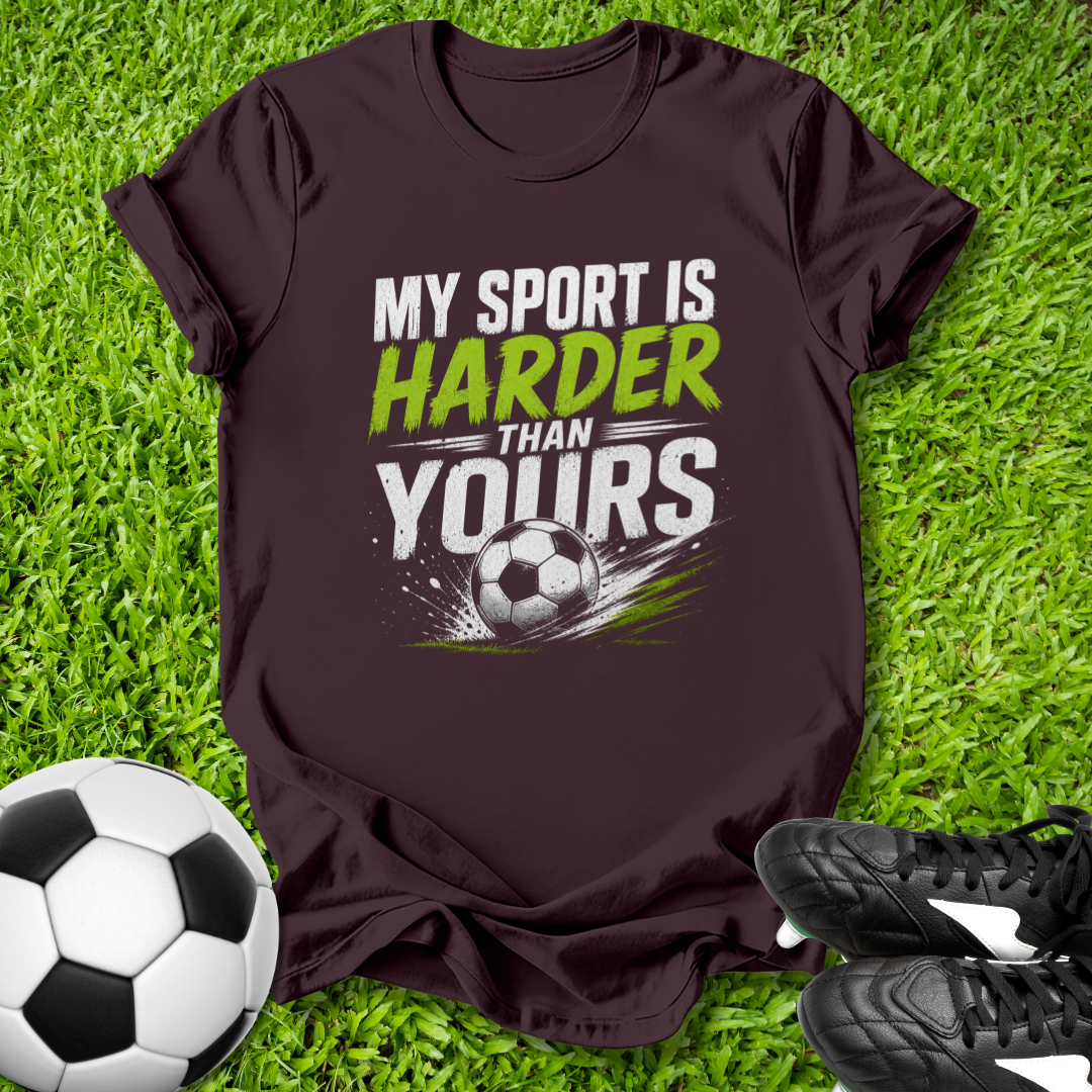 Maroon t-shirt with 'My sport is harder than yours' text on grass with soccer ball and shoes.