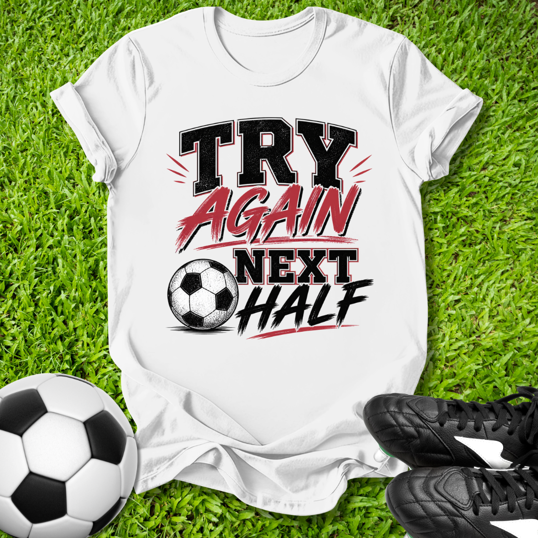 White t-shirt with 'Try Again Next Half' text on grass with soccer ball and shoes
