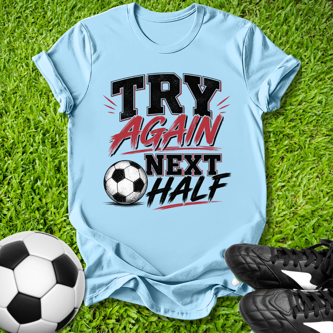 Light blue t-shirt with 'Try Again Next Half' text on grass with soccer ball and shoes.