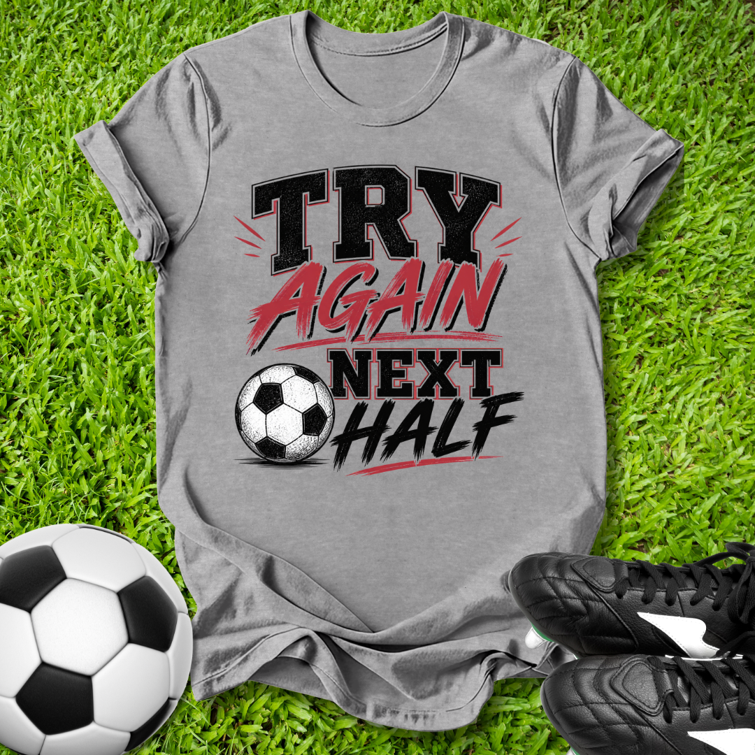 Gray t-shirt with 'Try Again Next Half' text on grass with soccer balls and shoes.
