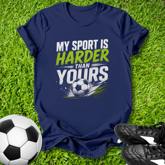 Navy blue t-shirt with 'My sport is harder than yours' text on grass with soccer ball and shoes.