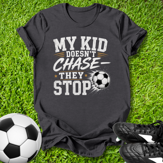 Black t-shirt with 'My kid doesn't chase, they stop' text on grass with soccer ball and shoes.