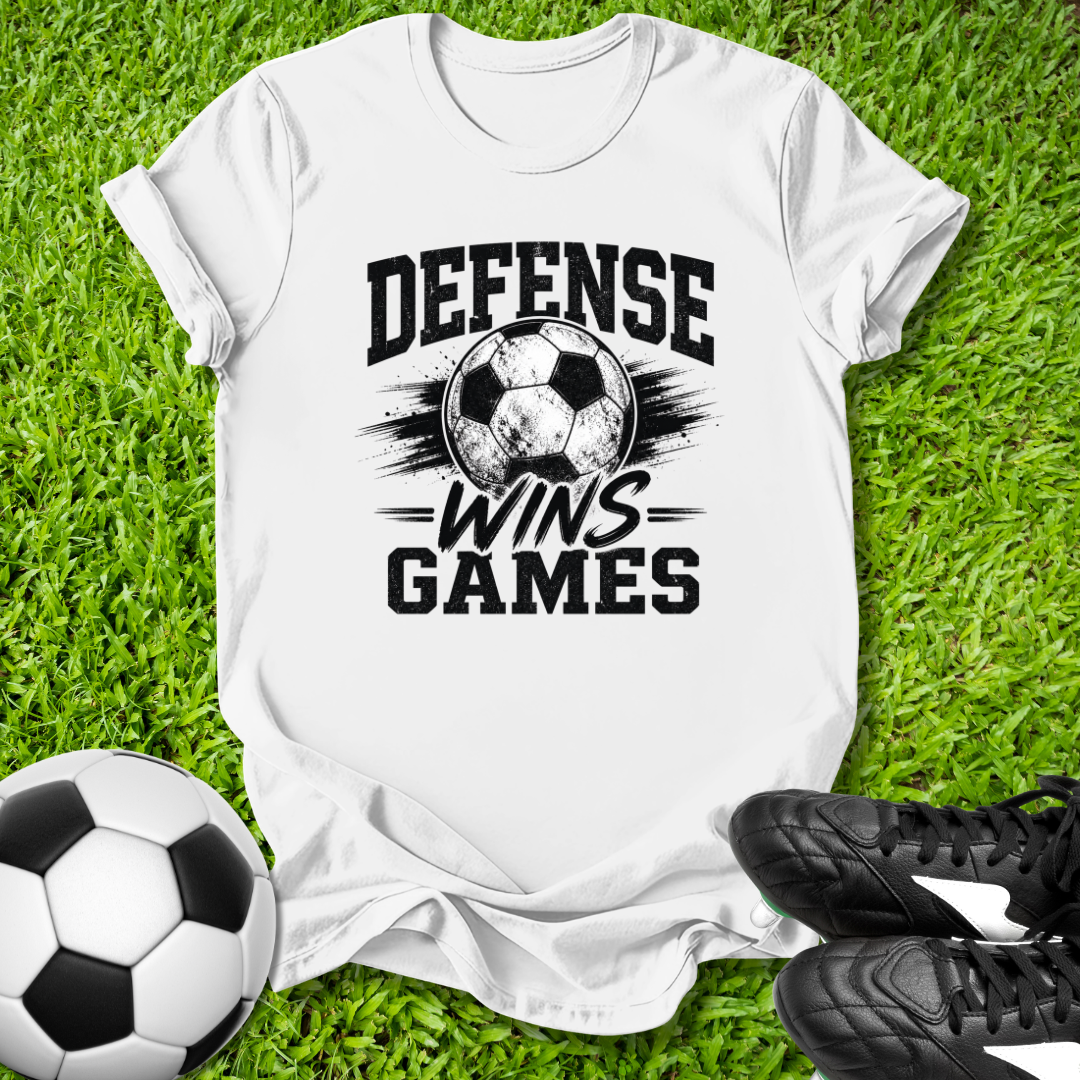 White t-shirt with 'Defense Wins Games' text on grass with soccer ball and shoes.