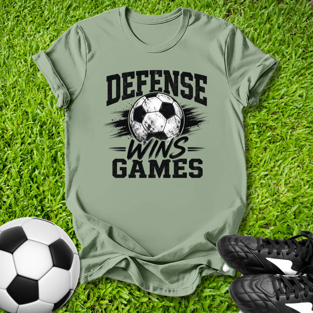 Green t-shirt with soccer ball and text 'Defense Wins Games' on grass with soccer ball and shoes.