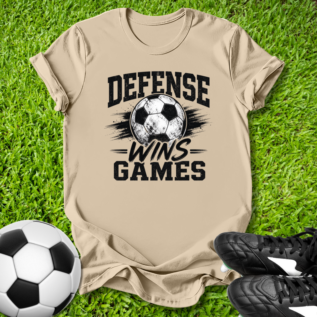 Beige t-shirt with soccer ball and text 'Defense Wins Games' on grass, with a soccer ball and cleats.