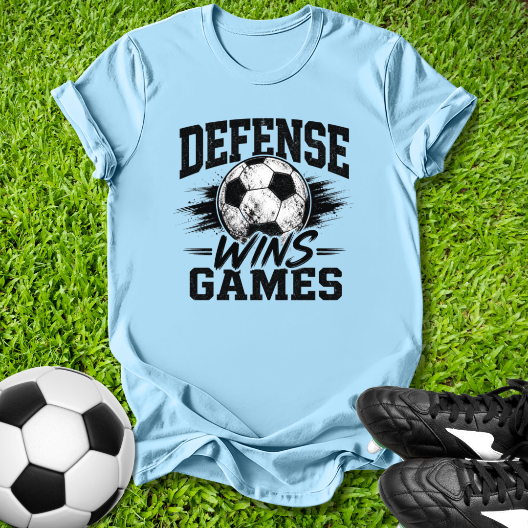 Light blue t-shirt with 'Defense Wins Games' text and soccer ball graphic on grass, with soccer ball and shoes.