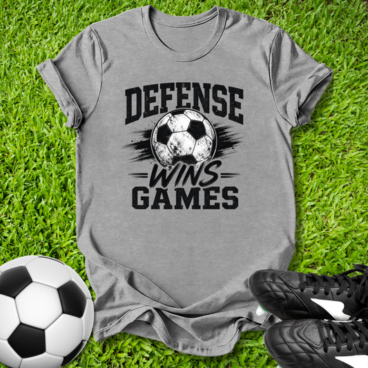 Gray -shirt with 'Defense Wins Games' text and soccer ball graphic on grass, with soccer ball and shoes.