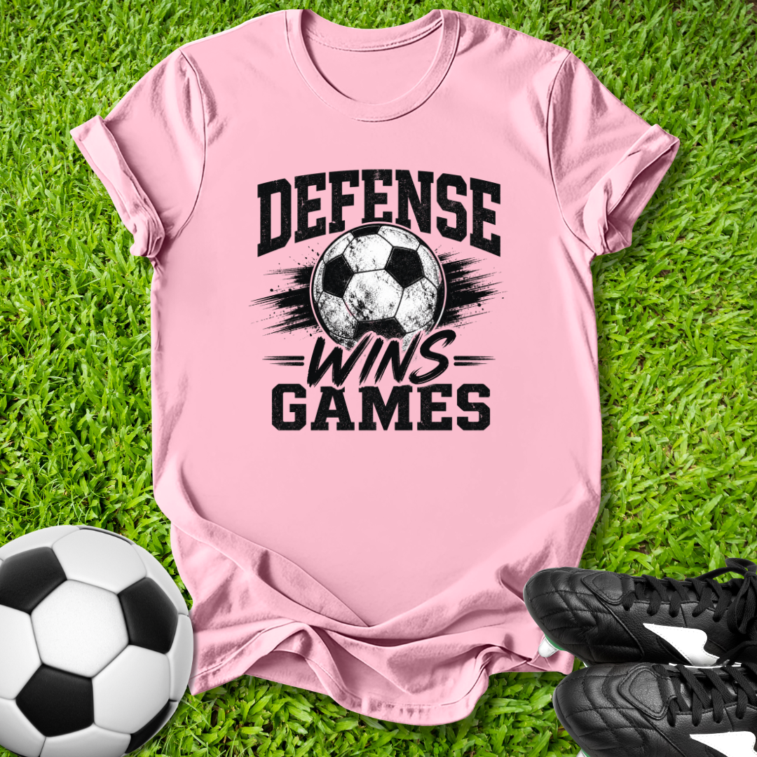 Pink t-shirt with 'Defense Wins Games' text and soccer ball on grass with soccer ball and shoes.