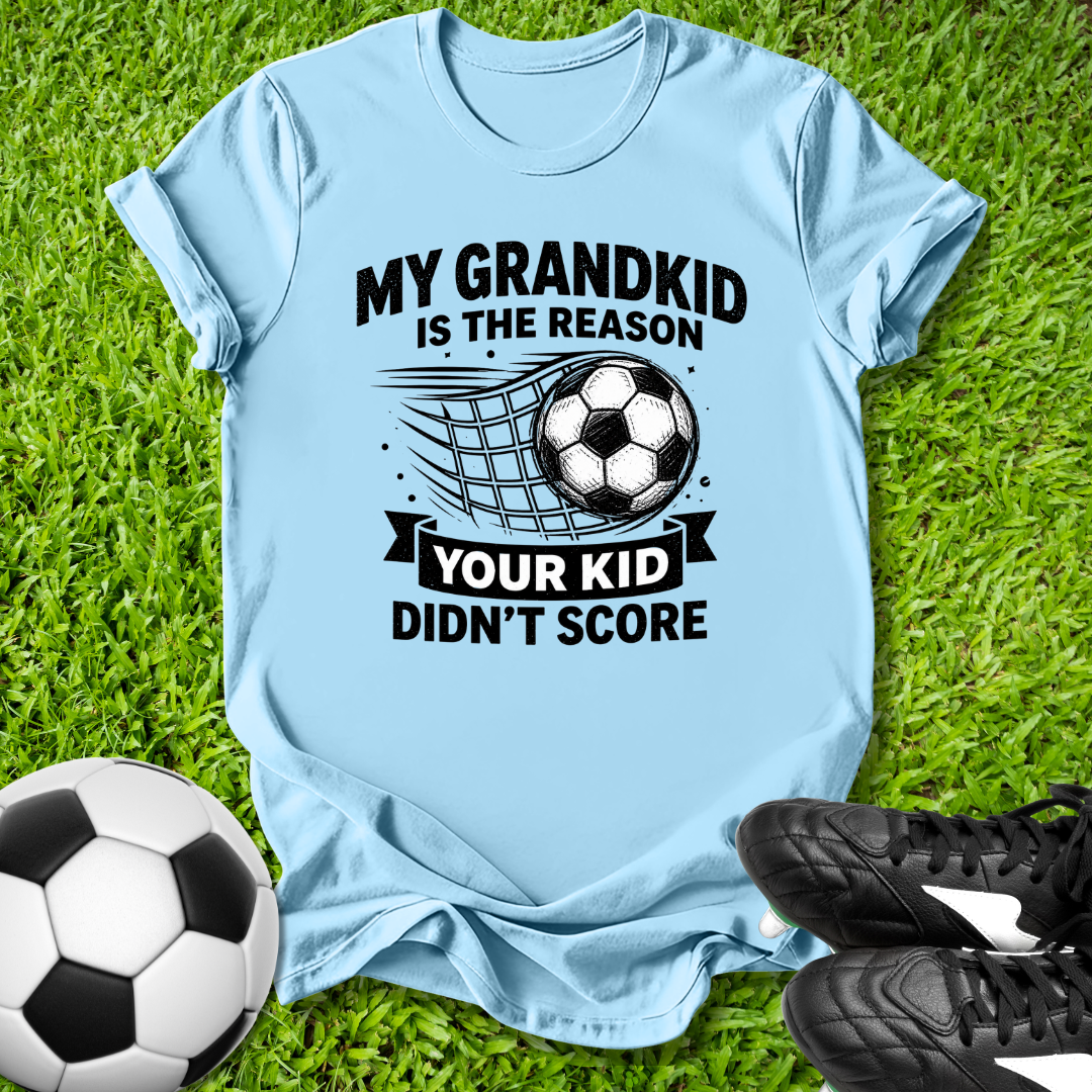 Light blue t-shirt with soccer-themed text on grass with soccer balls and shoes.